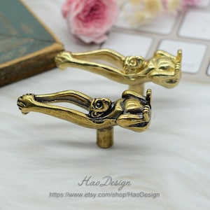 K&ouml;nnte beinhalten: Zwei goldfarbene Metall-Schrankkn&ouml;pfe in Form eines Affen mit ausgestreckten Armen. Der Affe sitzt mit gebeugten Beinen. Die Kn&ouml;pfe befinden sich auf einer wei&szlig;en Oberfl&auml;che.