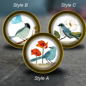 Könnte beinhalten: Drei runde, goldgerahmte, glasartige Ornamente mit verschiedenen Vogelillustrationen. Das erste Ornament zeigt einen blauen Vogel mit roten Mohnblumen, das zweite Ornament zeigt einen blauen Vogel, der eine Blase bläst, und das dritte Ornament zeigt einen blauen Vogel mit einer weißen Magnolienblüte.