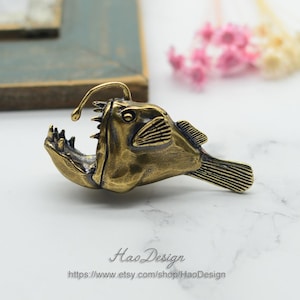 May include: A brass anglerfish figurine with a detailed design. The fish has a large mouth with sharp teeth and a lure on its head. The figurine is a unique and interesting piece of art.