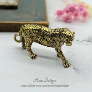 Pode incluir: Uma figurinha de tigre de metal dourado com listras detalhadas. A figurinha est&aacute; em p&eacute; sobre uma superf&iacute;cie branca.