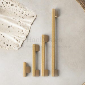 May include: Four gold-toned cabinet pulls of varying sizes, arranged vertically. The pulls have a textured, dotted pattern on the handle and a solid, rectangular base. A textured, off-white decorative element is in the upper left corner.