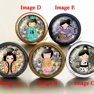 Pode incluir: Cinco botões redondos para armário com acabamento dourado, prateado e preto. Cada botão apresenta uma ilustração colorida de uma boneca japonesa vestindo um quimono e adereços tradicionais para o cabelo. As bonecas estão sobre um fundo de padrões florais e geométricos.