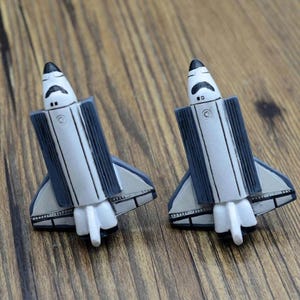May include: Two white and blue space shuttle figurines with black details. The figurines are facing forward and have a detailed design.