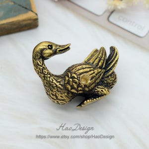 May include: A gold-coloured metal goose figurine with detailed textured wings and a curled neck. The goose is lying on its side with its head turned to the right. The figurine has a small hole on its back.