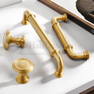 May include: A collection of gold-colored cabinet hardware, including two curved handles, a small knob, and a larger round knob. The hardware has a brushed finish and a classic design, suitable for furniture or cabinetry.