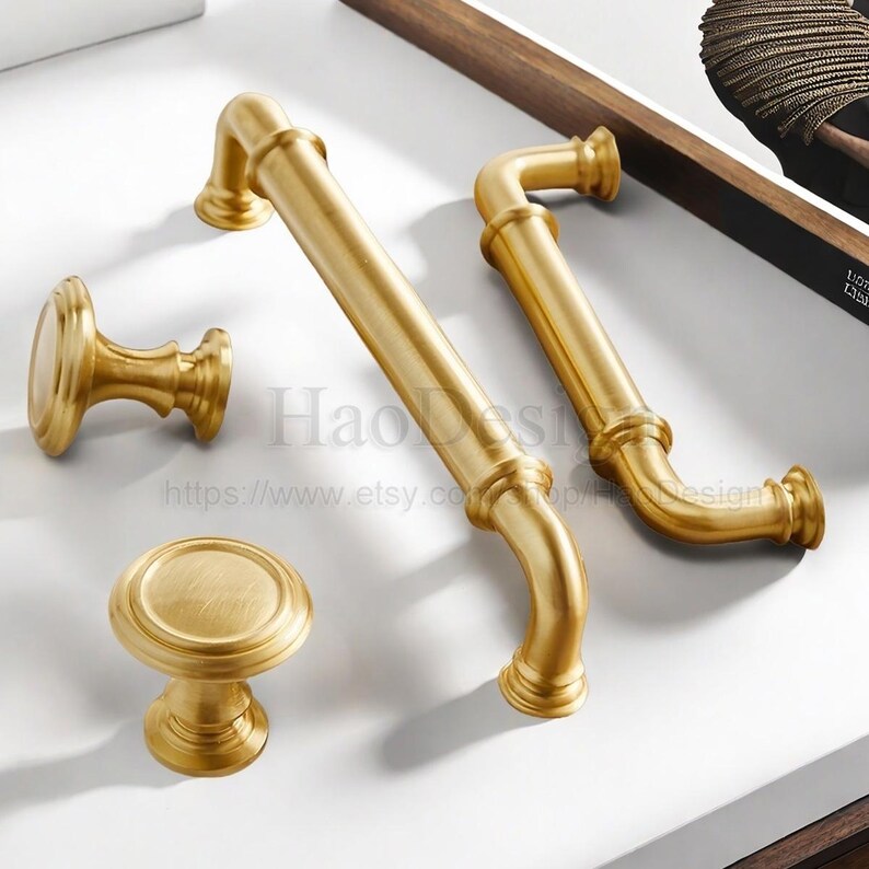 May include: A collection of gold-colored cabinet hardware, including two curved handles, a small knob, and a larger round knob. The hardware has a brushed finish and a classic design, suitable for furniture or cabinetry.