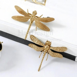 以下が含まれることがあります： 2つの金色のトンボの装飾品。 トンボは、詳細な羽と体を持っており、表面はテクスチャ加工されています。 トンボは白い表面に配置され、黒いストライプが見えます。