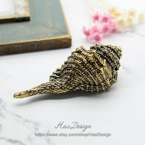 May include: A gold-colored seashell with a detailed, textured surface. The shell is a decorative object with a long, pointed end.