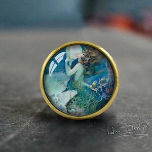 May include: A round, gold-toned, decorative knob with a glass insert featuring a vintage illustration of a mermaid with flowing hair, surrounded by bubbles and sea life.