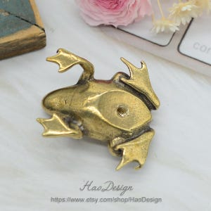 Może przedstawiać: Złota figurka żaby o szczeg&oacute;łowym wzorze. Żaba jest zwr&oacute;cona do g&oacute;ry, z rozłożonymi nogami. Figurka ma otw&oacute;r z tyłu do zawieszenia.