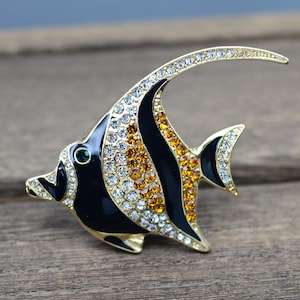 Op de afbeelding: Een broche van goudkleurig metaal in de vorm van een zwarte en oranje engelvis met heldere en amberkleurige strasssteentjes.