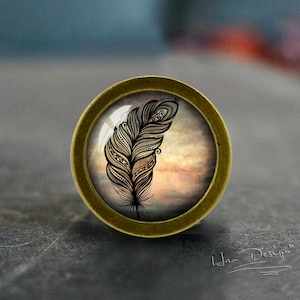 Pode incluir: Um botão redondo de cor latão com um inserto de vidro que apresenta um design de pena estilizada em preto e branco sobre um fundo de aquarela.