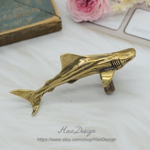 May include: A gold-coloured brass shark figurine with a detailed design. The shark is positioned in a swimming pose with its mouth open and its fins spread out. The figurine is a decorative piece and would be a great addition to any home decor.