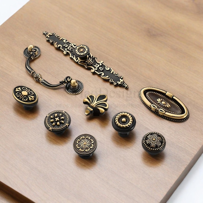 以下が含まれることがあります： アンティークスタイルのキャビネットハードウェアのコレクション。黒とゴールドの装飾的なデザインのノブとプルが含まれています。さまざまな形状とサイズがあり、明るい茶色の木製の表面に配置されています。