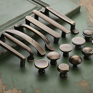 May include: A selection of bronze-coloured cabinet hardware, featuring different handle and knob designs. The handles are rectangular and curved, and the knobs are round with flat or rounded tops. The hardware is displayed on a distressed green surface.