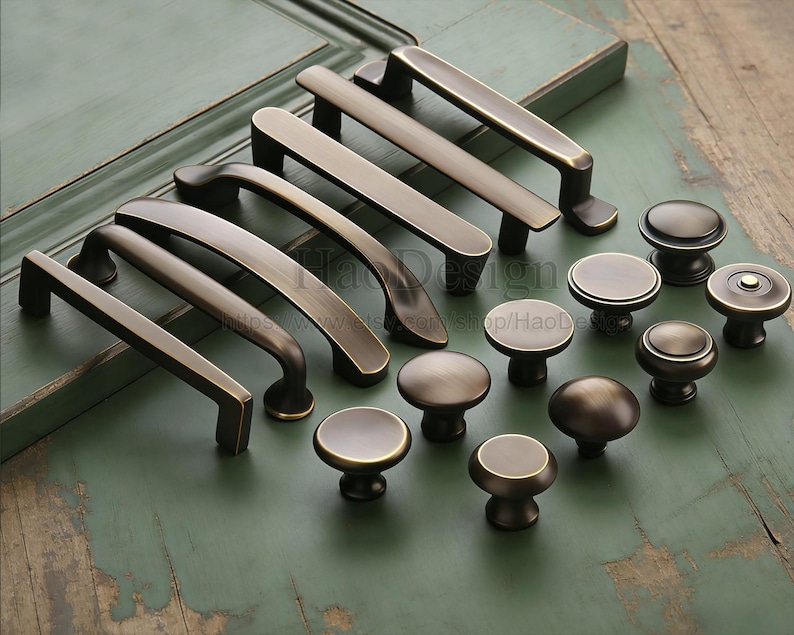May include: A selection of bronze-coloured cabinet hardware, featuring different handle and knob designs. The handles are rectangular and curved, and the knobs are round with flat or rounded tops. The hardware is displayed on a distressed green surface.