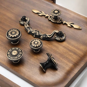 Puede incluir: Un conjunto de herrajes para armarios en negro y dorado, que incluye pomos y tiradores, dispuestos sobre una superficie de madera. Los pomos presentan un diseño floral, mientras que los tiradores tienen detalles ornamentados. Adecuado para muebles o armarios.