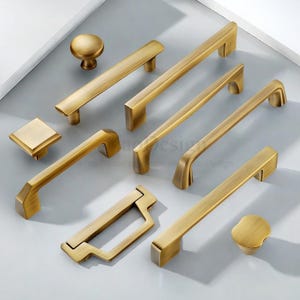 May include: A collection of antique brass cabinet hardware, including knobs and various handle styles. The hardware has a warm, aged finish, and is arranged on a light gray surface. The hardware is designed for use on cabinets and drawers.