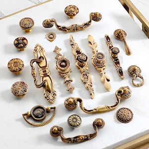 Op de afbeelding: Een verzameling antieke messing kastbeslagen, waaronder knoppen, handgrepen en decoratieve achterplaten. De hardware heeft sierlijke bloemen- en scrollwerkontwerpen in verschillende vormen en maten. De hardware wordt weergegeven op een wit oppervlak.