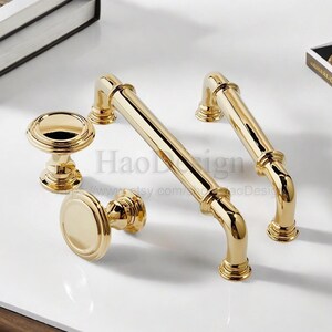 May include: A set of gold-colored cabinet hardware, including two bar pulls and two knobs. The hardware has a shiny finish and a classic design, suitable for drawers and cabinets. The hardware is made of metal.