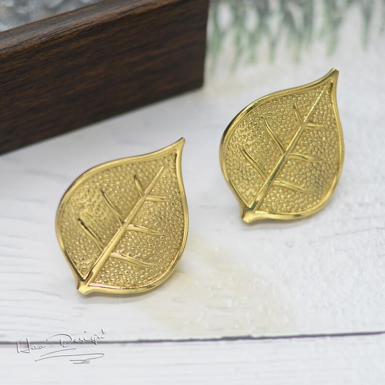 K&ouml;nnte beinhalten: Ein Paar goldene Ohrringe in Blattform. Die Ohrringe sind texturiert und haben ein detailliertes Blattdesign.