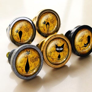 May include: Five round cabinet knobs with a gold, black, or silver finish. Each knob features a black silhouette of a cat on a yellow background with a swirling floral pattern.