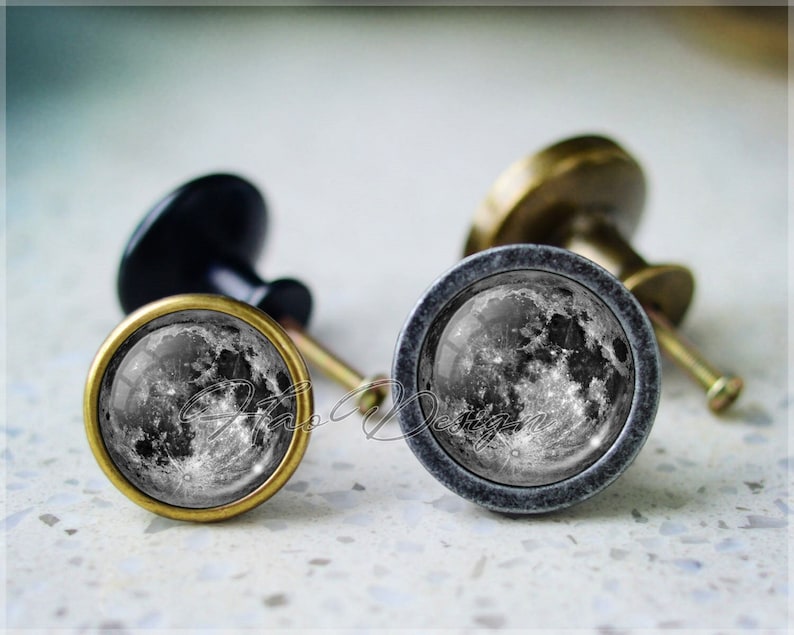 May include: Two cabinet knobs with a moon image. The knob on the left has a brass finish and the knob on the right has a silver finish.