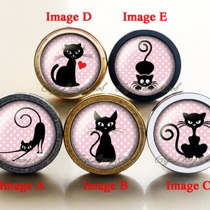 Könnte beinhalten: Fünf verschiedene schwarze Katzenmotive auf runden Schrankknöpfen. Jeder Knopf hat einen rosa Hintergrund mit weißen Punkten. Die Knöpfe sind aus Metall und haben eine glänzende Oberfläche.