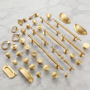 May include: A collection of gold-colored cabinet hardware, including knobs, pulls, and handles. Various shapes and sizes are displayed, from small round knobs to long, ornate handles. The hardware is arranged on a white and gray marble surface.