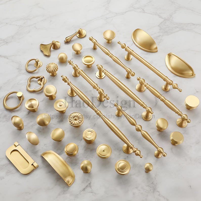May include: A collection of gold-colored cabinet hardware, including knobs, pulls, and handles. Various shapes and sizes are displayed, from small round knobs to long, ornate handles. The hardware is arranged on a white and gray marble surface.