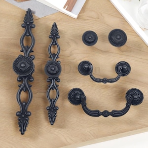 Może przedstawiać: Zestaw czarnych okuć do szafek, w tym dwa długie uchwyty, dwa gałki i dwa uchwyty wiszące. Okucia charakteryzują się ozdobnymi, inspirowanymi kwiatami wzorami i są ułożone na jasnej drewnianej powierzchni.