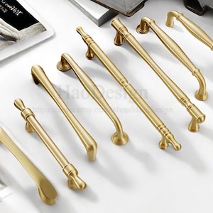 May include: A collection of gold-colored cabinet handles in various designs. The handles are arranged on a white surface, showcasing their different shapes and sizes. Some have curved designs, while others are straight with decorative ends.