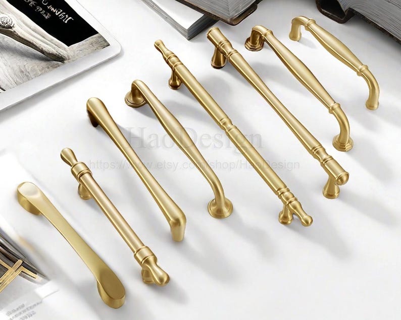 May include: A collection of gold-colored cabinet handles in various designs. The handles are arranged on a white surface, showcasing their different shapes and sizes. Some have curved designs, while others are straight with decorative ends.