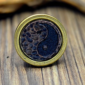 Puede incluir: Un colgante redondo de color latón con un símbolo yin yang negro y blanco en el centro. El símbolo es texturizado y tiene un acabado martillado.