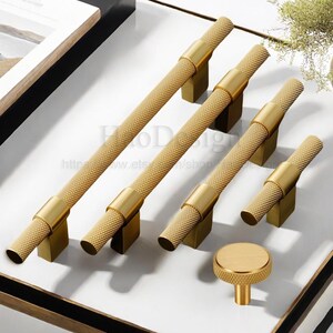 May include: A collection of gold-toned cabinet hardware, including several bar pulls and a round knob. The pulls have a textured, diamond-patterned grip and are mounted on square bases. The hardware is displayed on a white surface.