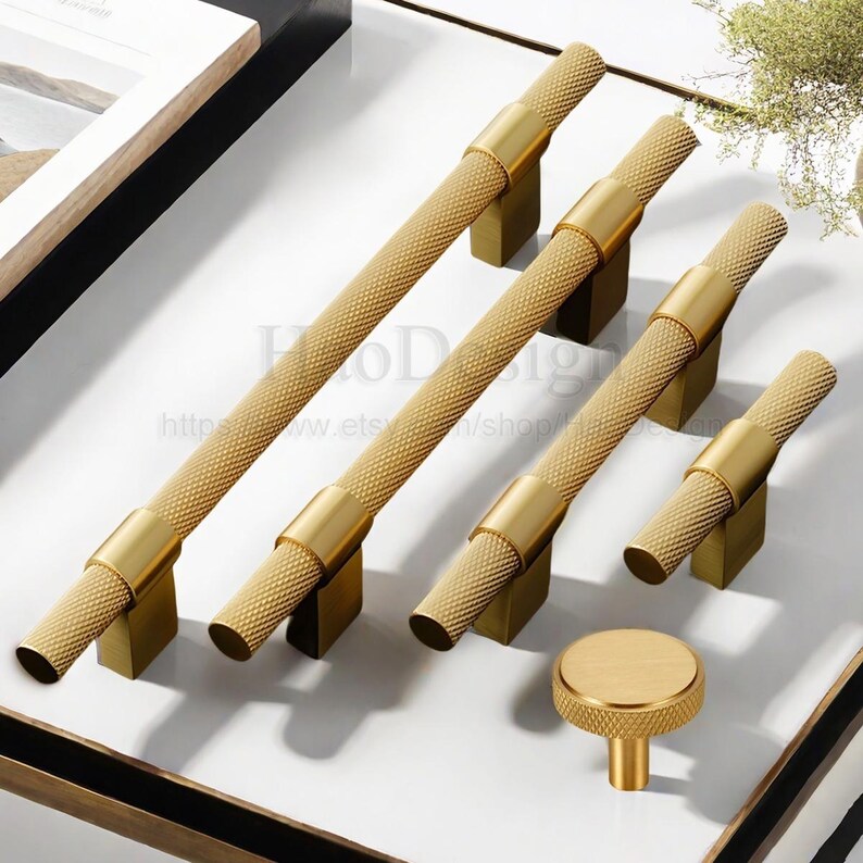 May include: A collection of gold-toned cabinet hardware, including several bar pulls and a round knob. The pulls have a textured, diamond-patterned grip and are mounted on square bases. The hardware is displayed on a white surface.