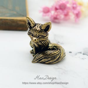 Puede incluir: Una pequeña figurita de metal de color dorado de un zorro sentado con la cola enroscada alrededor de su cuerpo. El zorro tiene una superficie detallada y texturizada que se asemeja al pelaje.