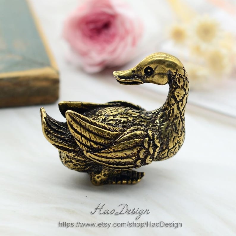 May include: A small, gold-coloured metal duck figurine with intricate details. The duck is sitting with its head turned to the side and its wings spread out.