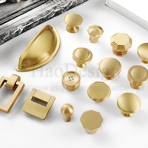 May include: A collection of gold-toned cabinet hardware, including knobs and pulls, in various shapes and sizes. The hardware includes round, octagonal, and rectangular designs. The hardware is arranged on a white surface.