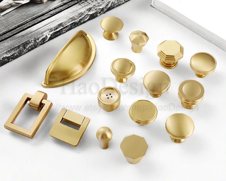 May include: A collection of gold-toned cabinet hardware, including knobs and pulls, in various shapes and sizes. The hardware includes round, octagonal, and rectangular designs. The hardware is arranged on a white surface.