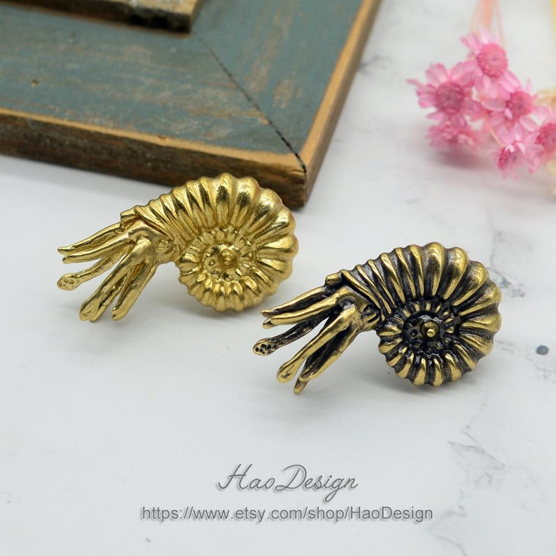 May include: A pair of gold-toned ammonite earrings with a detailed, textured design. The earrings are a unique and stylish accessory for any occasion.