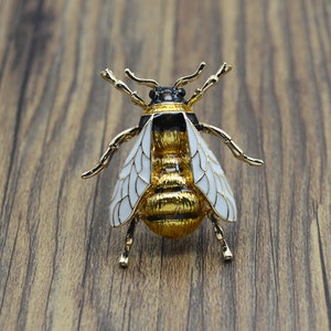 Op de afbeelding: Een gouden bij broche met witte vleugels en zwarte accenten. De bij is gedetailleerd met een textuur lichaam en heeft een pin aan de achterkant.