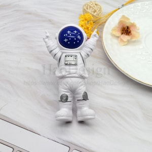 May include: A white astronaut figurine with arms raised, featuring a blue helmet with white star accents. The astronaut has silver details and is standing on a white surface. The figurine is a decorative item.