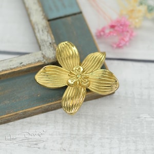 Pode incluir: Uma flor de metal dourada com cinco pétalos e um detalhe no centro. A flor está sobre uma superfície de madeira clara.