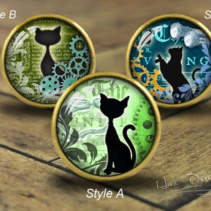 Könnte beinhalten: Drei runde, dekorative Glascabochons mit einer Messingfassung. Jeder Cabochon zeigt eine schwarze Silhouette einer Katze auf einem anderen Hintergrund. Der erste Cabochon hat einen grün-blauen Hintergrund mit einem Zahnradmuster. Der zweite Cabochon hat einen grün-weißen Hintergrund mit einem Blumenmuster. Der dritte Cabochon hat einen blau-goldenen Hintergrund mit einem Blumenmuster.