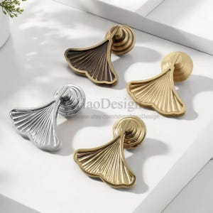 May include: Four cabinet pulls in silver, gold, and bronze finishes. Each pull has a fan-shaped design with detailed veining, mounted on a circular base. The pulls are arranged on a white surface. Suitable for furniture.
