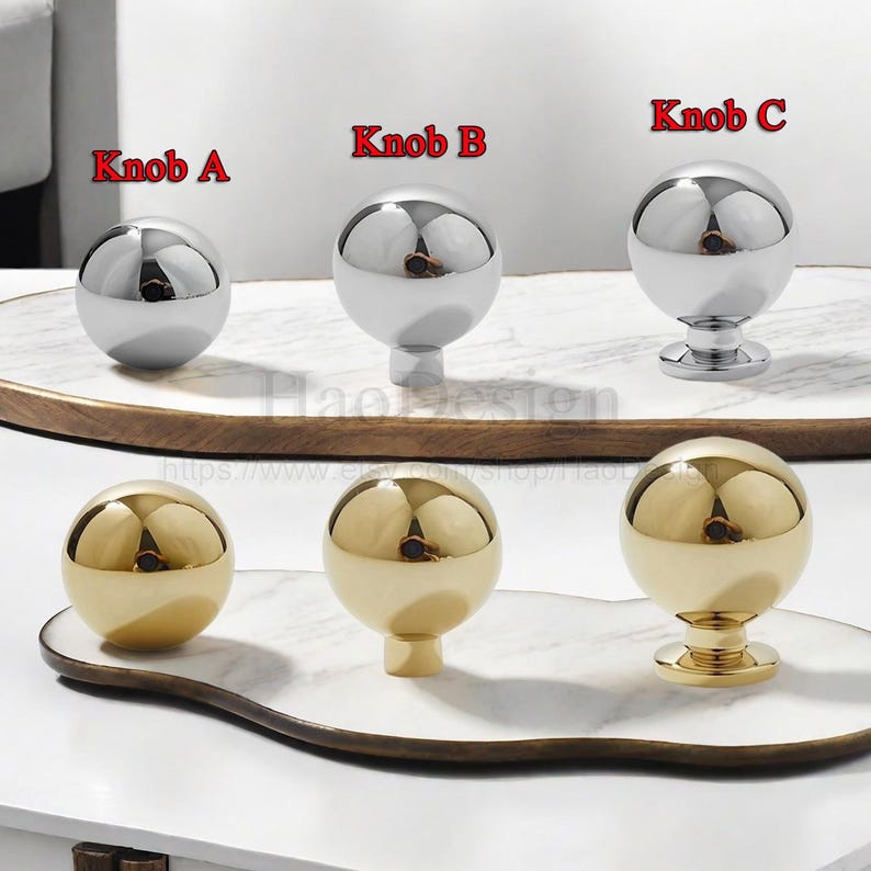 K&ouml;nnte beinhalten: Eine Sammlung von kugelf&ouml;rmigen Schrankkn&ouml;pfen in Silber- und Goldausf&uuml;hrungen. Die Kn&ouml;pfe sind mit "Knob A", "Knob B" und "Knob C" beschriftet. Die Kn&ouml;pfe werden auf einer wei&szlig;en und braunen Oberfl&auml;che pr&auml;sentiert.