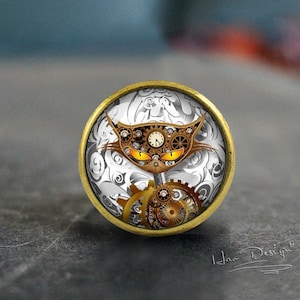 Peut inclure: Un bouton décoratif rond de couleur laiton avec un fond blanc et gris. Le bouton présente une illustration détaillée d'un visage de chat fait de rouages et d'un cadran d'horloge. Les yeux du chat sont orange vif.
