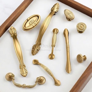 K&ouml;nnte beinhalten: Eine Sammlung von antiken Messing-Schrankbeschl&auml;gen, darunter Griffe und Kn&ouml;pfe. Die Beschl&auml;ge weisen verzierte Blumen- und Blattmuster mit einem warmen, goldenen Finish auf. Die Teile sind auf einer wei&szlig;en Oberfl&auml;che in einem Holzrahmen angeordnet. Ma&szlig;e in cm.