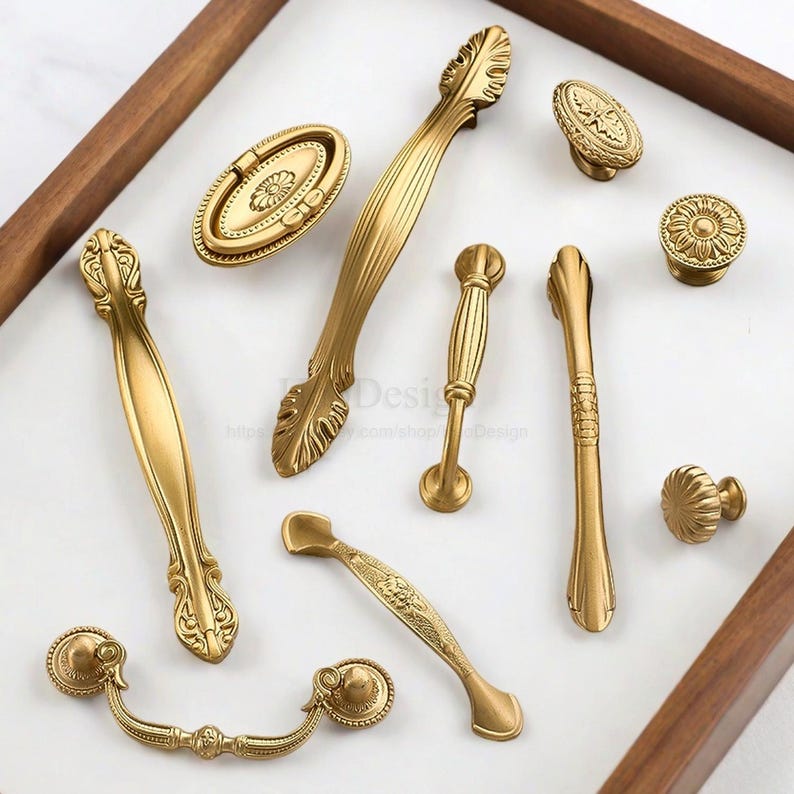 K&ouml;nnte beinhalten: Eine Sammlung von antiken Messing-Schrankbeschl&auml;gen, darunter Griffe und Kn&ouml;pfe. Die Beschl&auml;ge weisen verzierte Blumen- und Blattmuster mit einem warmen, goldenen Finish auf. Die Teile sind auf einer wei&szlig;en Oberfl&auml;che in einem Holzrahmen angeordnet. Ma&szlig;e in cm.